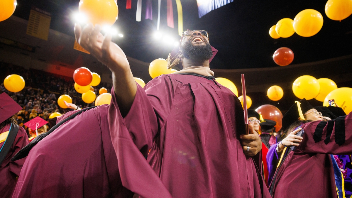 Black man with beard and glasses wearing maroon cap and gown looks up at balloons dropping during commencement